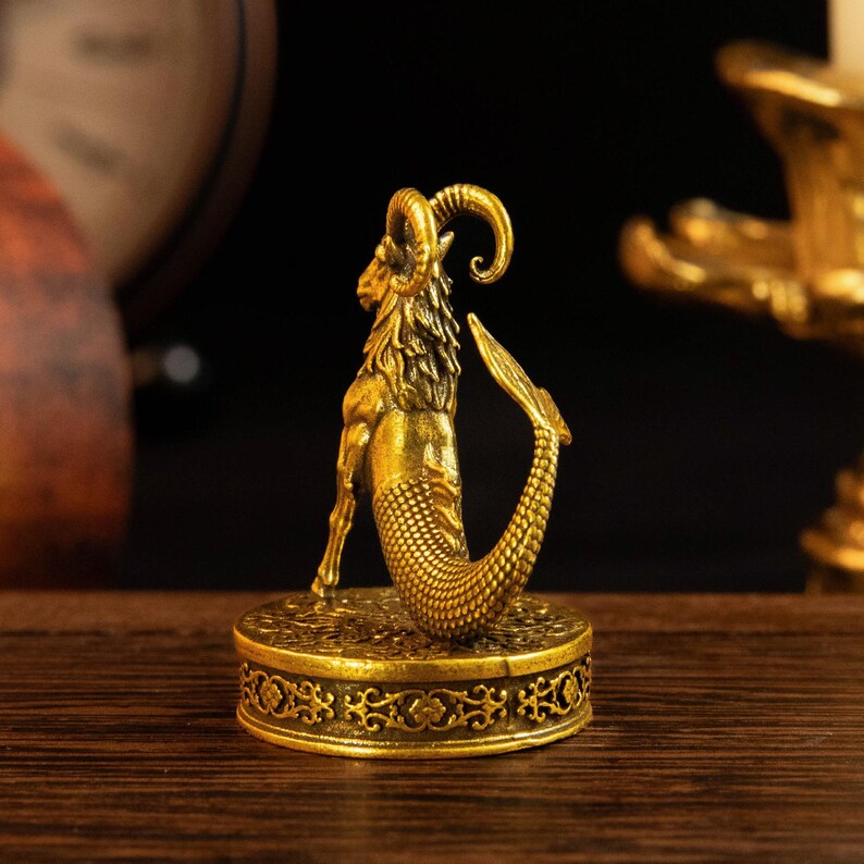 May include: A gold-colored brass figurine of a Capricorn sea-goat, featuring the upper body of a goat with curved horns and the lower body of a fish with scales. The creature stands on a round, ornate base decorated with intricate floral patterns.