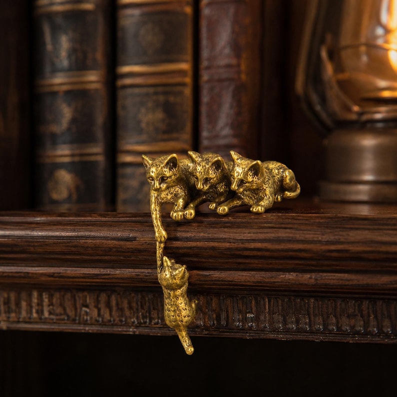 May include: A gold-toned metal figurine featuring four cats. Three cats are positioned on a horizontal surface, while one cat hangs vertically from the paw of one of the cats on the ledge. The cats have textured surfaces and detailed features.