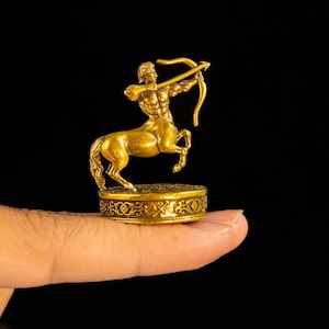 May include: A small, gold-colored metal figurine of a centaur archer. The centaur is depicted in a dynamic pose, drawing a bow with an arrow. The figure stands on a circular, ornate base featuring intricate, embossed scrollwork patterns.