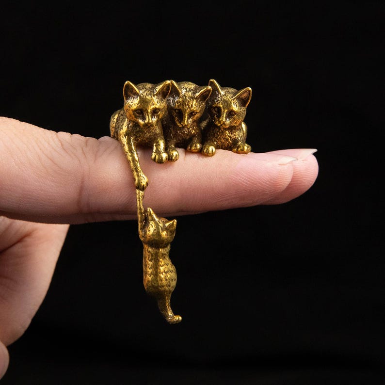May include: A gold-toned metal ring featuring a cluster of three small cat figures sitting on top, with a fourth cat figure hanging down from the paw of one of the top cats. The metal has a textured, antique finish with dark recessed details.