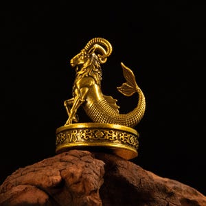 May include: A gold-colored metal figurine of a Capricorn sea-goat with a goat head and a fish tail. The creature stands on a cylindrical base featuring an intricate filigree pattern and the Capricorn zodiac symbol engraved on the front side.
