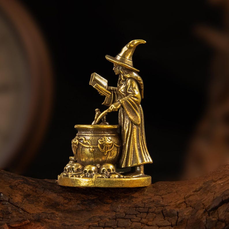 Puede incluir: Una figura de metal color lat&oacute;n que representa a un personaje con t&uacute;nica y sombrero puntiagudo, de pie junto a un caldero. El personaje sostiene un libro abierto con una mano y remueve el caldero con la otra. La base est&aacute; decorada con calaveras.