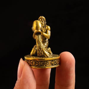 May include: A small, metallic gold-colored figurine depicting a kneeling figure pouring water from a large vessel. The figure is mounted on a circular base featuring intricate embossed floral patterns and the Aquarius zodiac symbol.