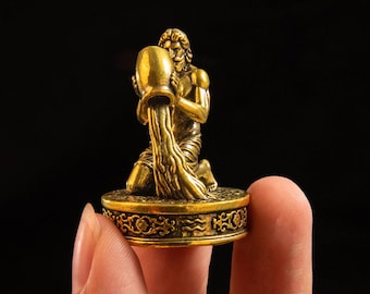 May include: A small, metallic gold-colored figurine depicting a kneeling figure pouring water from a large vessel. The figure is mounted on a circular base featuring intricate embossed floral patterns and the Aquarius zodiac symbol.