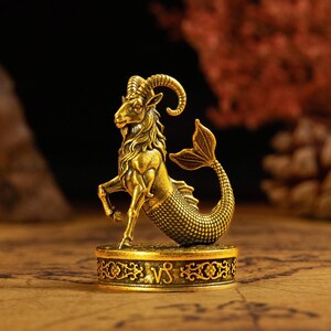 May include: A gold-toned metal figurine of a Capricorn zodiac creature, featuring the upper body of a goat with curved horns and the lower body of a fish with scales. The figure stands on a circular base engraved with the Capricorn symbol.