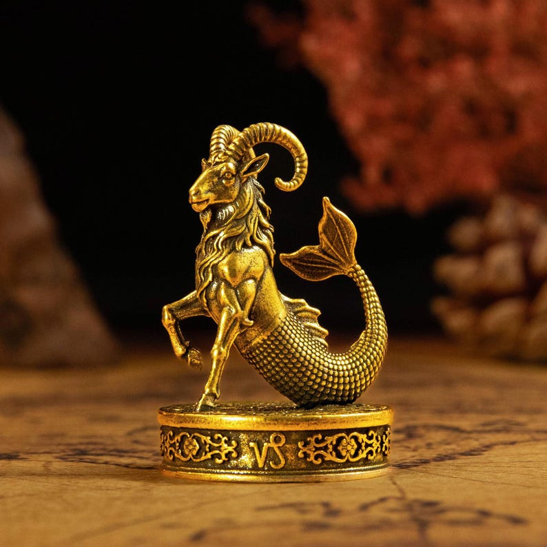 May include: A gold-toned metal figurine of a Capricorn zodiac creature, featuring the upper body of a goat with curved horns and the lower body of a fish with scales. The figure stands on a circular base engraved with the Capricorn symbol.