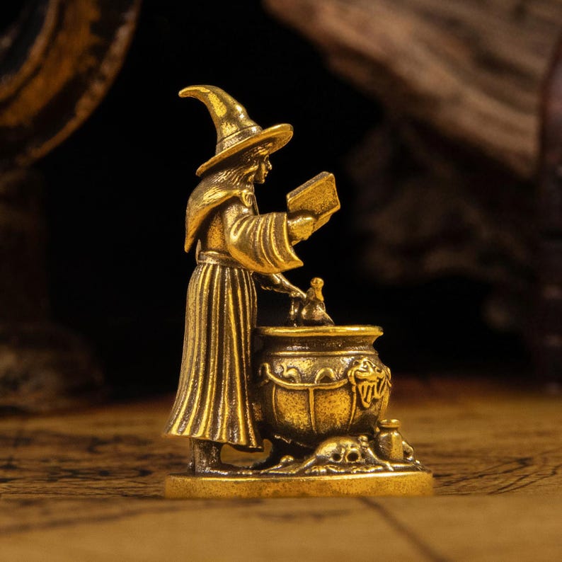 Puede incluir: Una figura de metal en tono dorado que representa a un personaje con t&uacute;nica y sombrero puntiagudo, de pie junto a un caldero grande. El personaje sostiene un libro abierto en una mano y alcanza el caldero, que presenta detalles de calaveras.