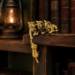 May include: A gold-toned metal figurine featuring four cats. Three cats are positioned on a horizontal surface, while one cat hangs vertically from the paw of one of the cats above, creating a connected chain of feline figures with textured details.