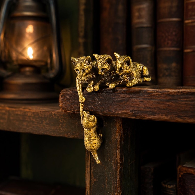 May include: A gold-toned metal figurine featuring four cats. Three cats are positioned on a horizontal surface, while one cat hangs vertically from the paw of one of the cats above, creating a connected chain of feline figures with textured details.