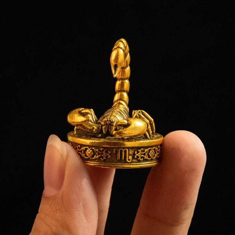 May include: A small, metallic gold-colored scorpion figurine mounted on a circular base. The base features an engraved Scorpio zodiac symbol and intricate scrollwork patterns. The scorpion is depicted with its tail arched upward and claws extended.