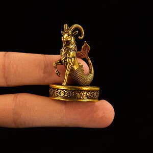 May include: A small, metallic gold-colored figurine of a Capricorn sea-goat with curved horns and a scaled fish tail. The creature stands on a circular base featuring an intricate filigree pattern and the engraved zodiac symbol for Capricorn.