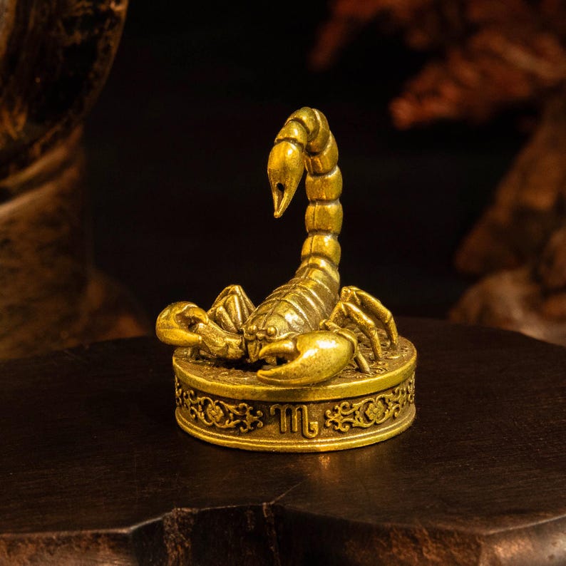 May include: A small, metallic gold-colored scorpion figurine perched atop a circular base. The base features an intricate floral border and the Scorpio zodiac symbol. The scorpion is depicted with its tail arched upward and claws extended forward.