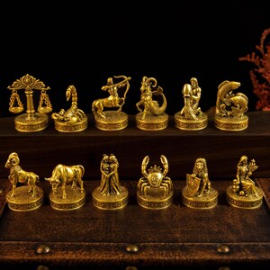 May include: A collection of twelve gold-toned metal zodiac figurines, each mounted on a circular base with embossed detailing. The set includes representations of Libra, Scorpio, Sagittarius, Capricorn, Aquarius, Pisces, Aries, Taurus, Gemini, Cancer, Leo, and Virgo.
