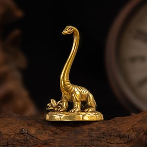 May include: A small, polished brass figurine of a long-necked dinosaur standing on a textured, circular base. The dinosaur has a smooth, metallic finish with visible anatomical details on its body and a small plant-like element at its feet.