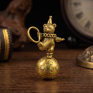 May include: A brass figurine of a bear dressed as a clown, wearing a pointed hat and a ruffled collar. The bear is balancing on one foot atop a spherical ball decorated with embossed stars, while holding a circular hoop in one raised hand.