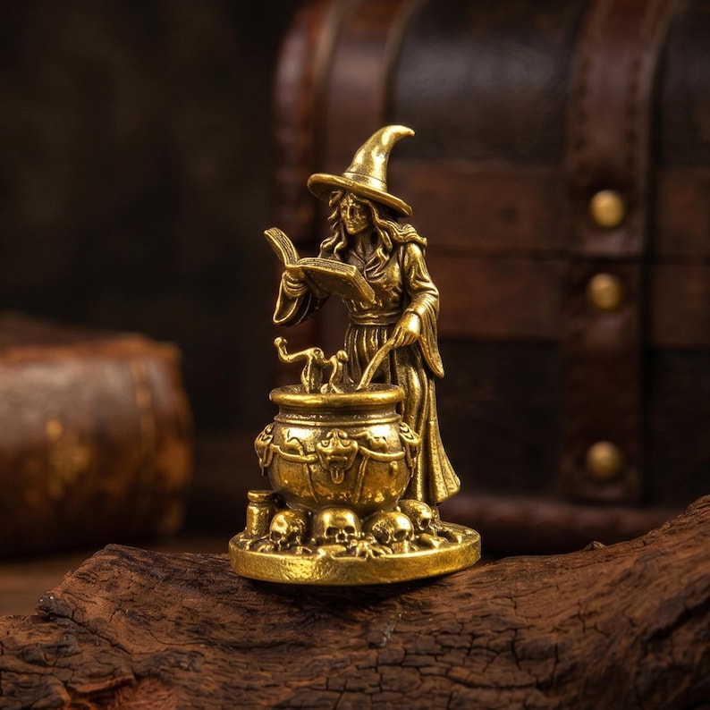 Puede incluir: Una figura de metal color lat&oacute;n que representa a un personaje con t&uacute;nica y sombrero puntiagudo, de pie junto a un caldero adornado con motivos de calaveras. El personaje sostiene un libro abierto en una mano y un agitador en la otra, sobre una base.
