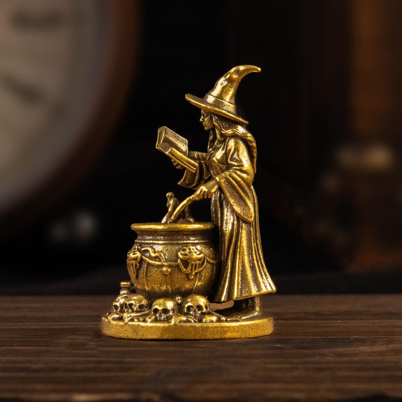 Puede incluir: Una figura de metal color lat&oacute;n que representa a un personaje con t&uacute;nica y sombrero puntiagudo, de pie junto a un caldero mientras sostiene un libro abierto. La base de la escultura est&aacute; decorada con varias calaveras peque&ntilde;as alrededor.