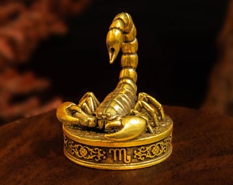 May include: A gold-toned metal scorpion figurine perched atop a circular base. The base features an intricate embossed floral pattern and the Scorpio zodiac symbol. The scorpion has a segmented tail curved upward and two large front pincers.