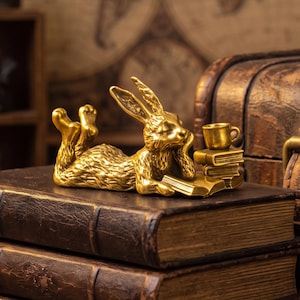 May include: A metallic gold-coloured rabbit figurine lying on its stomach, resting its head on one paw while reading an open book. Beside the rabbit are two stacked books and a small cup. The rabbit has textured fur and long ears.