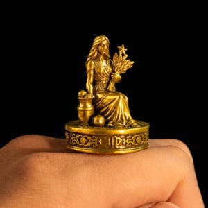 May include: A gold-toned metal ring featuring a sculpted figure of a seated person holding a sheaf of wheat and a star, positioned next to a small vase. The circular base is decorated with ornate scrollwork and the Virgo zodiac symbol.