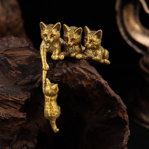 May include: A brass-colored metal figurine featuring four small cats. Three cats are positioned side-by-side on a ledge, while one cat hangs downward, held by the paw of one of the cats above. The metal has a textured, antique-style finish.