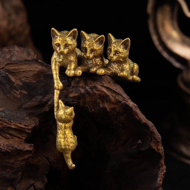 May include: A brass-colored metal figurine featuring four small cats. Three cats are positioned side-by-side on a ledge, while one cat hangs downward, held by the paw of one of the cats above. The metal has a textured, antique-style finish.