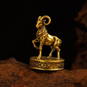May include: A metallic gold-coloured figurine of a ram standing on a circular base. The ram is depicted with curved horns and textured fur. The base features an intricate embossed floral pattern around its side and the Aries zodiac symbol.