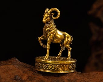May include: A metallic gold-colored figurine of a ram standing on a circular base. The ram is depicted with curved horns and textured fur. The base features an intricate embossed floral pattern around its side and the Aries zodiac symbol.