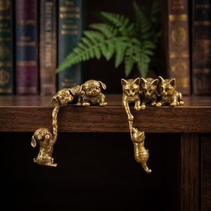 May include: A set of two brass-colored metal figurines. One figurine features two puppies, with one puppy hanging from the edge by the paw of the other. The second figurine features three kittens, with one kitten hanging from the edge by the paw of another.