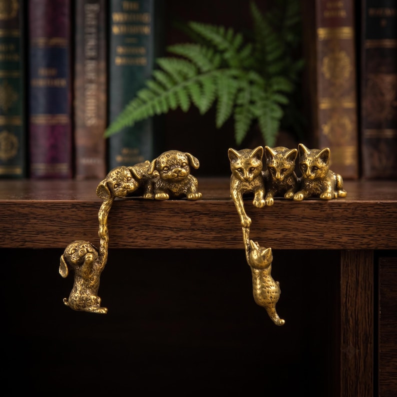 May include: A set of two brass-colored metal figurines. One figurine features two puppies, with one puppy hanging from the edge by the paw of the other. The second figurine features three kittens, with one kitten hanging from the edge by the paw of another.
