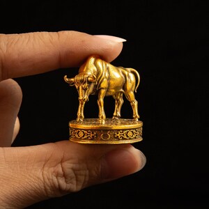 May include: A small gold-colored metal figurine of a bull standing on a circular base. The base features intricate scrollwork patterns and a small Taurus zodiac symbol engraved on the front side. The bull is depicted in a standing position.