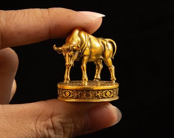 May include: A small gold-colored metal figurine of a bull standing on a circular base. The base features intricate scrollwork patterns and a small Taurus zodiac symbol engraved on the front side. The bull is depicted in a standing position.