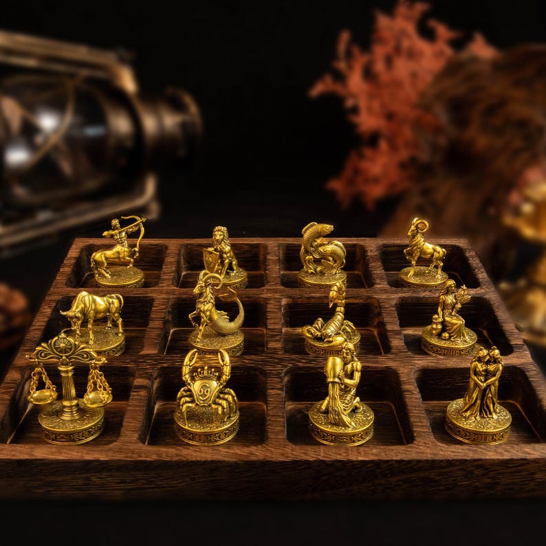 May include: A set of twelve gold-toned metal zodiac figurines arranged in a dark brown wooden grid tray. Each small statue represents a different astrological sign, including figures like a crab, a bull, a centaur, and a scale, all with detailed textures.