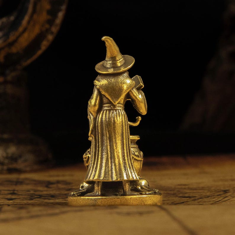 Puede incluir: Una peque&ntilde;a figura de color lat&oacute;n que representa a un personaje con sombrero puntiagudo y t&uacute;nica larga, de pie detr&aacute;s de un caldero. El personaje sostiene un objeto rectangular en una mano, sobre una base circular plana.