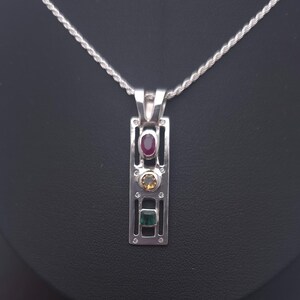 May include: A silver pendant necklace featuring a rectangular openwork frame set with three vertically aligned gemstones: a top oval red ruby, a middle round yellow citrine, and a bottom square green emerald, suspended on a silver rope chain.
