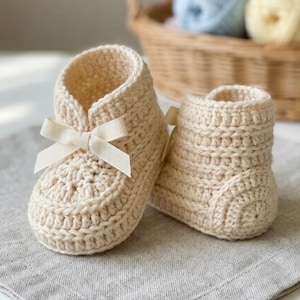 May include: A pair of cream-coloured crochet booties featuring a textured stitch pattern and a small, matching cream ribbon bow tied at the front of the ankle opening. The booties are crafted from yarn and have a high-top design with a rounded toe.