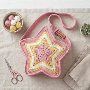 May include: A star-shaped crochet bag featuring concentric layers of pink, yellow, and white yarn. The bag includes a long shoulder strap with a small rectangular wooden tag embossed with the word HANDMADE attached to the strap hardware.