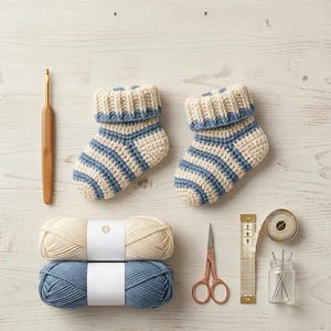 Könnte beinhalten: Ein Paar cremefarbene und blau gestreifte gehäkelte Babyschuhe, eine Häkelnadel aus Holz, zwei Garnknäuel, eine kupferfarbene Schere, ein Maßband und ein Glas mit Nadeln. Das Maßband zeigt Markierungen in Zentimetern an.