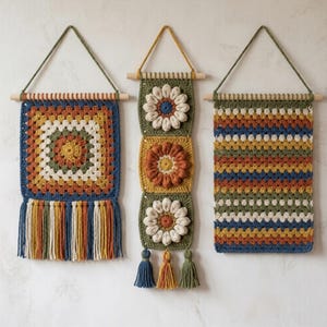 May include: Three handmade crochet wall hangings suspended from wooden dowels. The left piece features a square granny motif with fringe. The centre piece displays three stacked floral squares with tassels. The right piece has a striped pattern.