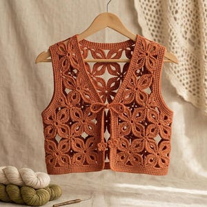 May include: A rust-colored crochet vest featuring an intricate openwork floral pattern throughout. The garment has a V-neckline, sleeveless armholes, and a front tie closure with small flower-shaped accents at the ends of the strings.