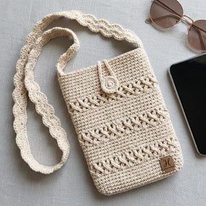 May include: A cream-coloured crochet crossbody bag featuring a scalloped strap, a button closure, and a small square leather tag with the letter H and the word handmade printed on it. The bag has a textured pattern with horizontal woven details.