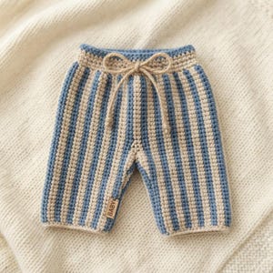 May include: Crocheted trousers featuring vertical stripes in light blue and cream. The waistband includes a tan drawstring tied in a bow at the centre. A small rectangular tag with the word BABY is attached to the lower left leg of the garment.