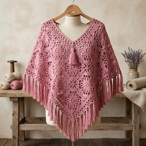 May include: A dusty rose crochet poncho featuring a V-neckline, a central tassel, and a repeating floral motif pattern. The garment is constructed from interconnected square panels and is finished with long, dangling fringe along the bottom edge.