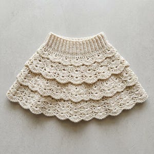 May include: A cream-colored crochet skirt featuring a ribbed waistband and three distinct, layered tiers with a scalloped edge design. The garment is crafted from yarn in a neutral off-white shade, showcasing a repetitive shell stitch pattern.