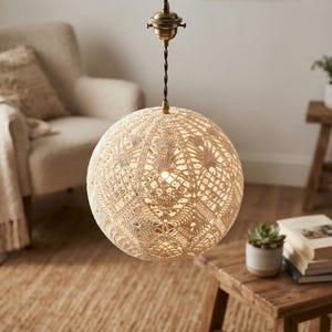 Op de afbeelding: Een bolvormige hanglamp met een gedetailleerde, crèmekleurige gehaakte kanten kap. De lamp hangt aan een gedraaid donker snoer aan een messingkleurige plafondbevestiging en verspreidt een warme gloed door de stof met het patroon.