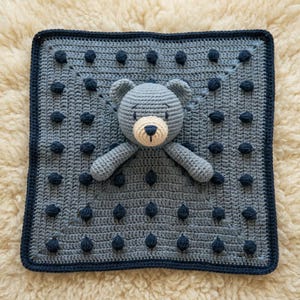 May include: A square crochet security blanket in light gray featuring a dark navy blue border and a grid of dark navy blue bobble stitches. Centered on the blanket is a stuffed crochet bear head and arms in light gray with a cream muzzle.