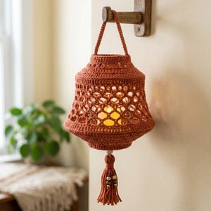 May include: A burnt orange crochet lantern cover featuring an intricate openwork lattice pattern. The lantern has a matching crochet handle and a decorative tassel at the bottom adorned with wooden beads, hanging from a wooden wall hook.