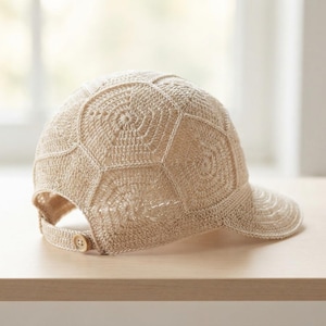 May include: A beige baseball cap constructed from crocheted yarn in a geometric hexagonal pattern. The cap features a curved brim and an adjustable back strap secured with a single light brown button, showcasing a detailed textile texture.