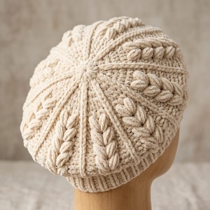 May include: A cream-coloured beret crafted from soft, plush velvet yarn. The hat features a radial design with alternating sections of smooth knit and raised, braided cable patterns that converge at the centre, finished with a ribbed knit brim.