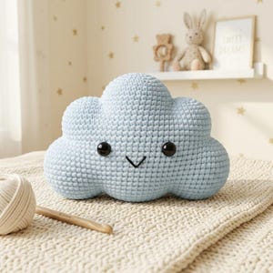 May include: A light blue cloud-shaped plush toy made of crocheted yarn. The toy features two round black plastic eyes and a simple black stitched V-shaped mouth. The texture is consistent with hand-knitted or crocheted fabric construction.